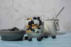 准备一步一步早....格兰诺拉麦片早餐自制的希腊酸奶蓝莓眼镜蓝色的木背景健康的饮食脆脆的格兰诺拉麦片酸奶坚果蓝莓