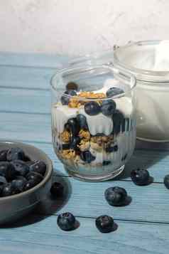 准备一步一步早....格兰诺拉麦片早餐自制的希腊酸奶蓝莓眼镜蓝色的木背景健康的饮食脆脆的格兰诺拉麦片酸奶坚果蓝莓