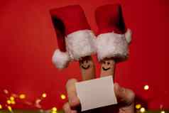 手指穿着圣诞老人的红白帽子快乐手指表情符号脸持有空纸请注意文本复制空间红色的背景快乐家庭庆祝概念圣诞节年一天夫妇