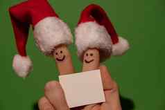 手指穿着圣诞老人的红白帽子快乐手指表情符号脸持有空纸请注意文本复制空间绿色背景快乐家庭庆祝概念圣诞节年一天夫妇