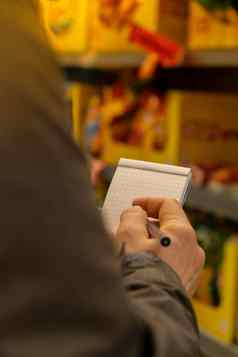 女手持有纸笔记本购物列表模糊货架上背景部分视图女人购物超市概念消费主义关闭女人使笔记购物列表