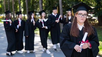 肖像年轻的高加索人女人眼镜研究生礼服背景同学们集团研究生学生在户外