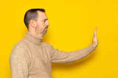 拒绝否认概念一边视图男人。穿高领毛衣使停止标志伸出的手警告表达式负手势室内工作室拍摄孤立的黄色的背景
