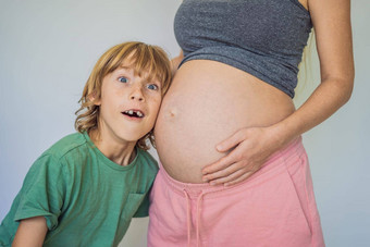 年轻的美丽的怀孕了女人老大儿子可爱的男孩靠耳朵母亲的肚子期待婴儿家庭概念准备老孩子年轻的