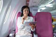 年轻的亚洲有吸引力的业务女人乘客坐着业务类奢侈品飞机穿无线耳机放松工作平板电脑飞行