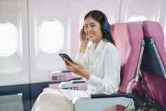 年轻的亚洲有吸引力的业务女人乘客坐着业务类奢侈品飞机穿无线耳机聪明的电话移动放松飞行