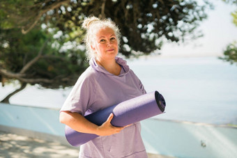 女人持有瑜伽席准备实践瑜伽冥想在户外海海滩快乐成熟的女人长发绺锻炼海滨冥想瑜伽放松概念