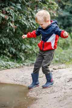 年轻的金发碧眼的男孩蓝色的红色的雨衣雨靴子幸福的水坑水泥