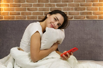 年轻的美丽的女人坐在床上拥抱枕头持有电话手相机