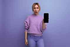 肖像惊讶金发碧眼的年轻的女人淡紫色毛衣智能手机模型明亮的背景复制空间
