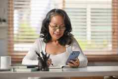 微笑成熟的女人坐着桌子上舒适的首页浏览移动应用程序聊天亲戚在线移动电话