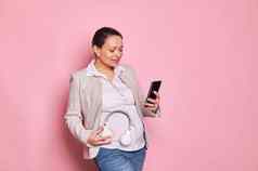 年轻的怀孕了女人连接孩子把音乐耳机肚子孤立的粉红色的背景