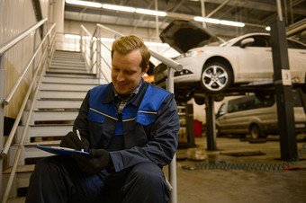 年轻的高加索人汽车机械师坐着楼梯修复商店写作检查清单剪贴板检查车车间机械维护工程师工作汽车行业
