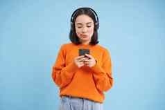 年轻的女人耳机智能手机听音乐享受唱歌最喜欢的歌曲播放列表蓝色的背景