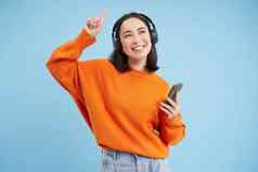 美丽的朝鲜文女人耳机跳舞听音乐移动电话应用程序微笑摆姿势蓝色的背景