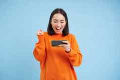 快乐朝鲜文女孩赢得移动电话视频游戏持有手机庆祝实现目标应用程序站蓝色的背景