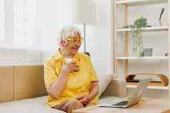 上了年纪的女人穿耳机移动PC沟通在线视频调用微笑幸福坐着沙发上首页工作黄色的衬衫生活方式退休女人