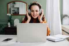 年轻的快乐女人学生穿耳机移动PC网络学习首页办公室听音乐生活讲座播客网络研讨会学习语言在线研讨会坐着桌子上电脑