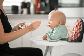 妈妈。勺子喂养婴儿男孩孩子婴儿椅子水果泥厨房首页婴儿固体食物介绍概念