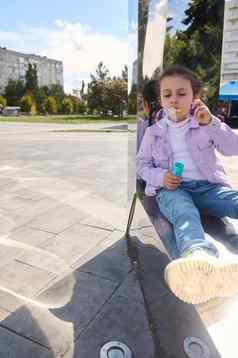 高加索人可爱的女孩休闲牛仔布坐着镜子金属板凳上吹肥皂泡沫城市公园