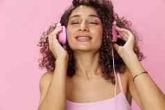 快乐女人穿耳机卷曲的头发听音乐唱歌眼睛关闭粉红色的t恤牛仔裤粉红色的背景复制空间