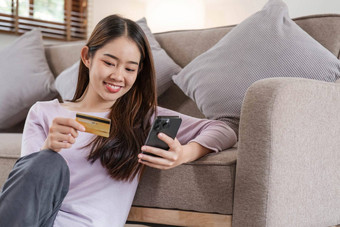 年轻的成人亚洲女消费者持有信贷卡智能手机坐着地板上首页在线银行事务商务虚拟购物安全移动银行概念