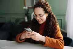 年轻的微笑女人穿眼镜持有智能手机手机现代技术移动检查细胞电话应用程序发短信浏览互联网购物坐着首页