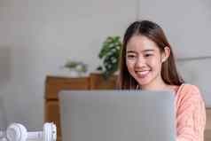 肖像微笑笑年轻的女人坐着表格穿无线耳机移动PC年轻的女看视频电影网络研讨会视频调用朋友家庭
