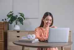 肖像微笑笑年轻的女人坐着表格穿无线耳机移动PC年轻的女看视频电影网络研讨会视频调用朋友家庭