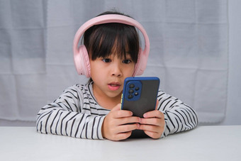 可爱的小学学校<strong>女孩</strong>穿耳机持有智能手机快乐亚洲<strong>女孩</strong>研究在线智能手机在家教育<strong>听音乐</strong>玩游戏