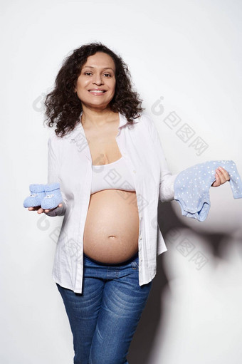 快乐的怀孕了女人持有蓝色的婴儿靴紧身衣裤未来新生儿孩子微笑白色背景