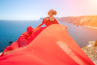 金发女郎长头发阳光明媚的海滨红色的流动衣服回来视图丝绸织物挥舞着风背景蓝色的天空山海滨