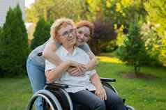高加索人女人拥抱上了年纪的妈妈。坐着轮椅走在户外