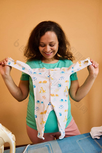 微笑怀孕了女人持有连身裤新生儿婴儿站熨衣服桌子上准备袋孕妇医院
