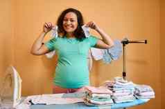 怀孕了女人持有新生儿衣服蒸汽铁微笑相机站熨衣服董事会熨婴儿衣服