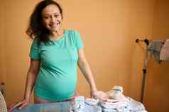 怀孕了女人积极的情感美丽的微笑脸站熨衣服董事会熨婴儿衣服