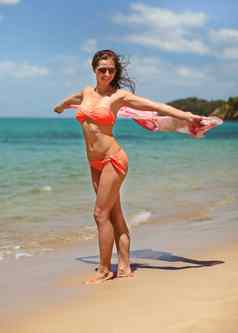 年轻的运动女人橙色比基尼泳装太阳镜走海滩持有围巾挥舞着风