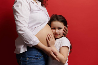 美丽的女孩拥抱肚子怀孕了妈妈。相机孤立的红色的背景