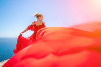 女人海红色的衣服金发女郎长头发阳光明媚的海滨红色的流动衣服回来视图丝绸织物挥舞着风背景蓝色的天空山海滨