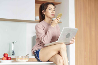 年轻的美丽的亚洲女性坐着厨房表格工作移动PC检查文本消息智能手机