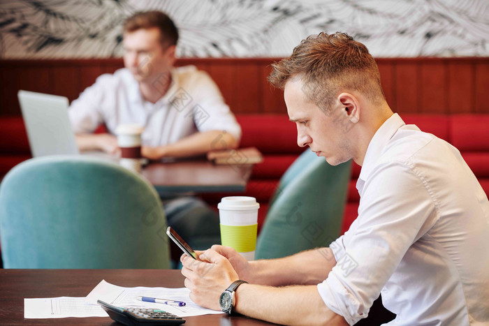 皱着眉头年轻的商人坐着咖啡馆表格文档前面阅读新闻智能手机屏幕