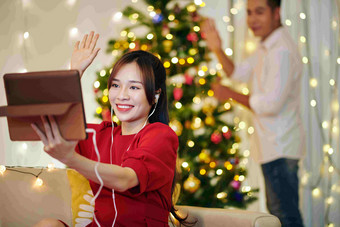 漂亮的微笑年轻的女人的男朋友挥舞着手视频调用朋友装修圣诞节树