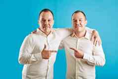 年轻的成人双胞胎兄弟站蓝色的墙背景指出手指相机微笑