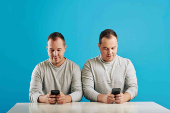水平拍摄双胞胎兄弟坐着表格看智能手机明亮的蓝色的背景