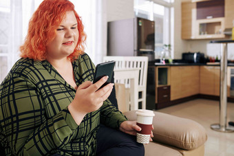 大小年轻的女人明亮的橙色头发坐着沙发首页喝咖啡检查消息智能手机