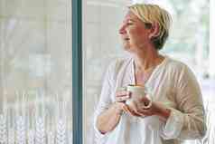 积极的成熟的女人白色棉花上衣喝杯子咖啡窗口