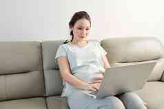 可爱的年轻的亚洲怀孕了女人休息沙发阅读文章移动PC屏幕