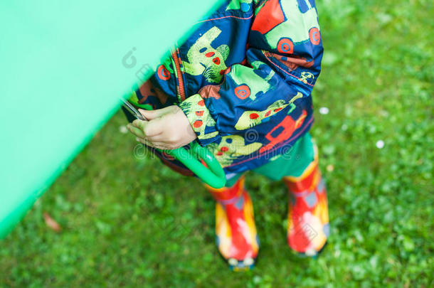 雨衣雨靴的小男孩躲在绿色雨伞下