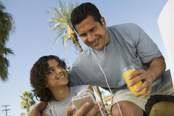 男孩（13-15岁），手持便携式<strong>音乐播放器</strong>，父亲戴着耳机听着<strong>音乐</strong>，手里拿着一杯果汁。