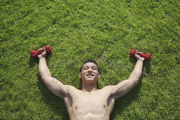在北京，赤膊上阵，微笑，肌肉发达的男子躺在草地上，举着哑铃，俯视